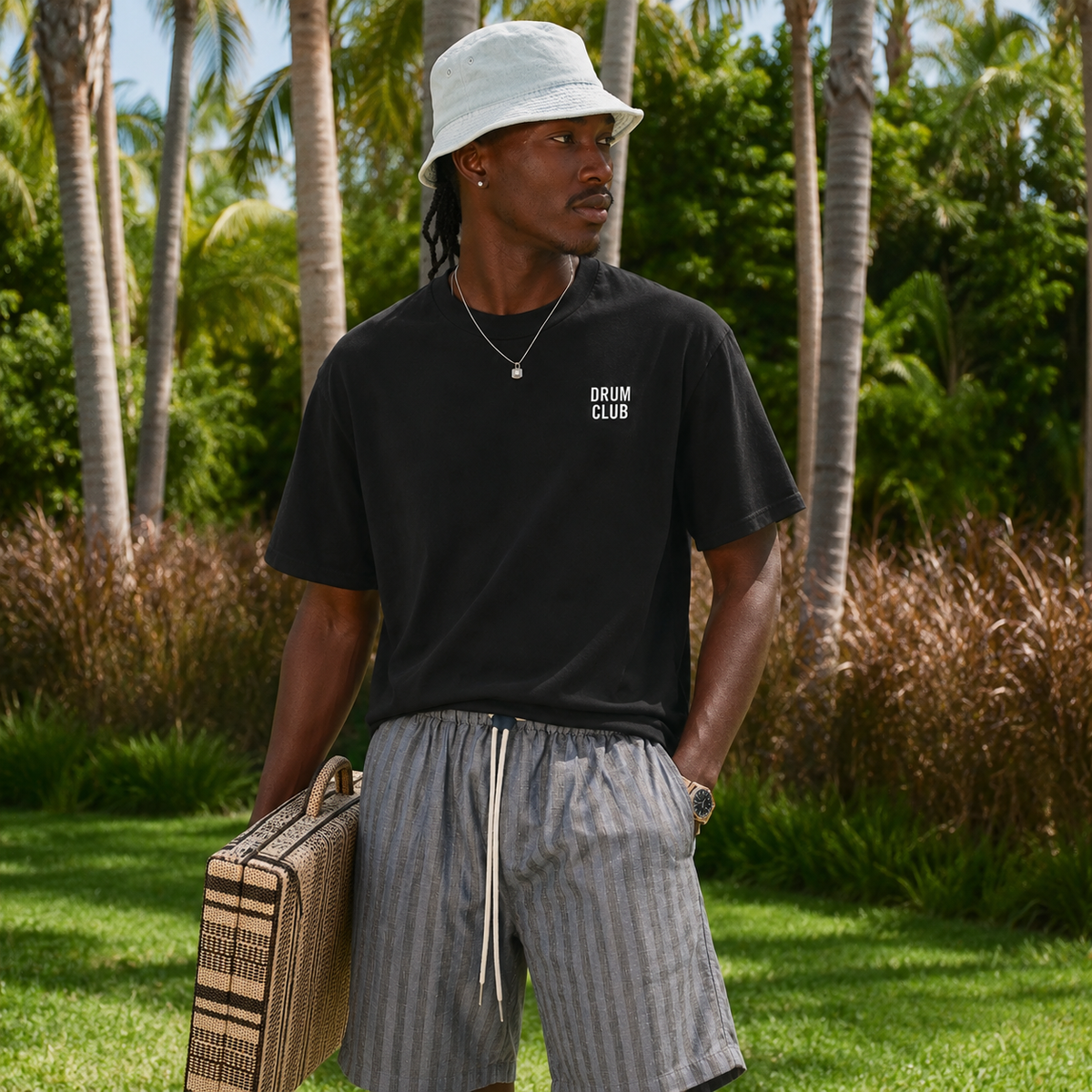 Male model wearing black DRUM CLUB t-shirt in tropical outdoor streetwear setting.