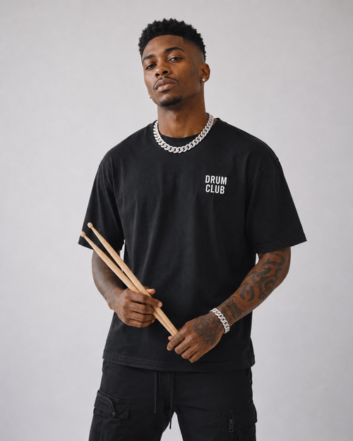 Black male model wearing a black DRUM CLUB T-shirt with a white left chest logo, holding drumsticks in a clean studio portrait.