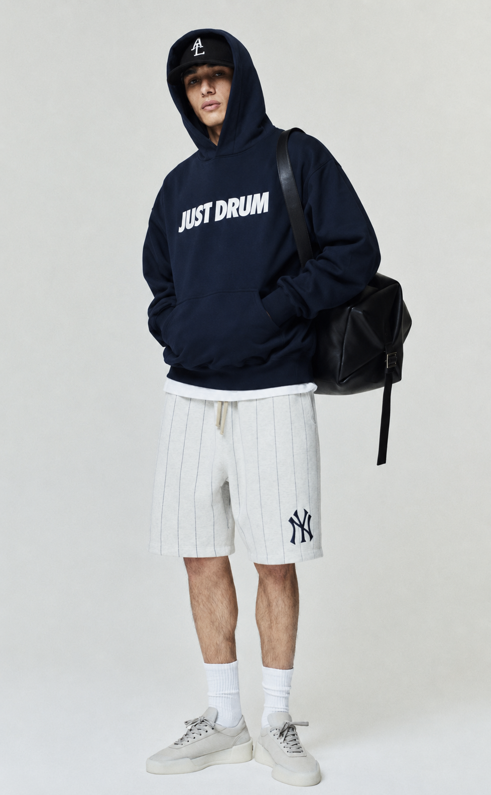 Person wearing a navy hoodie with 'JUST DRUM' text, white shorts with a logo, and carrying a black backpack on a light gray background.